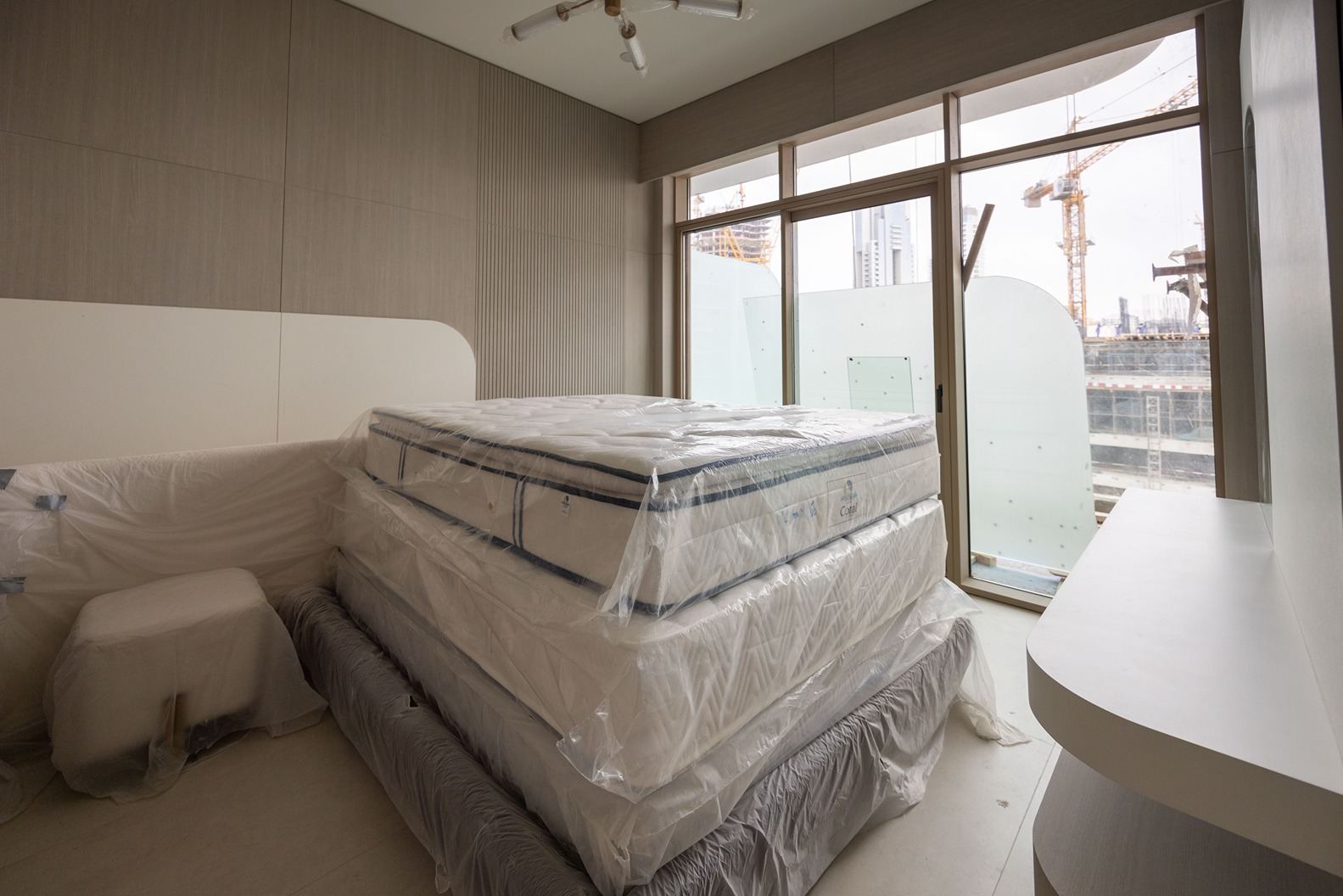Pearl House By Imtiaz A modern bedroom with stacked mattresses covered in plastic, large windows showing a construction site outside.